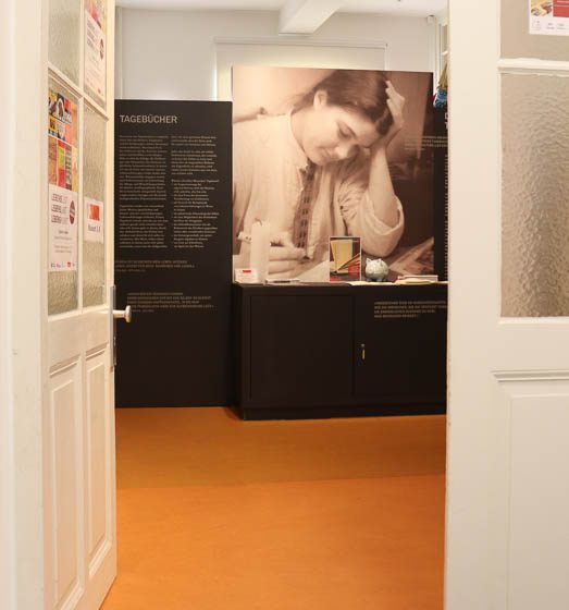 durch die geöffnete Tür des Museums blickt man auf das Foto einer Tagebuch schreibenden, jungen Frau über eine schwarzen Kommode und neben einer schwarzen Texttafel mit weißem Text