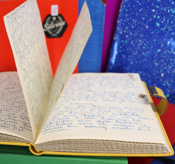 vor einem rot und blauen Hintergrund ein aufgeschlagenes, gelbes Buch, die Seiten dicht gefüllt mit handgeschriebenem Text